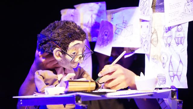 A puppet sits at a desk with drawings around them, you can see the puppeteers hand moving the head of the puppet