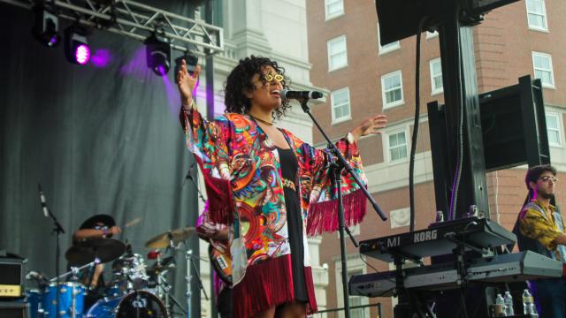 A woman in a colorful shawl sings with her hands outstretched on an outdoor stage.