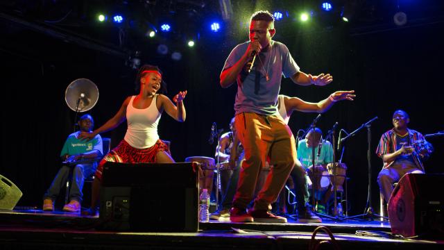 Jagwa Music performing in NYC, Photo by William Farrington People performing on stage, singing into a microphone and someone dancing in the background