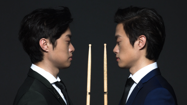 Twincussion by Terry Lin Two men facing each other in profile, each holding a drum stick