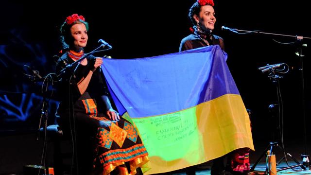 Two women sit on stage with microphones holding a Ukrainian flag between them