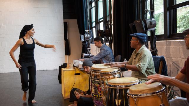 Woman in black pointing towards people playing the drums