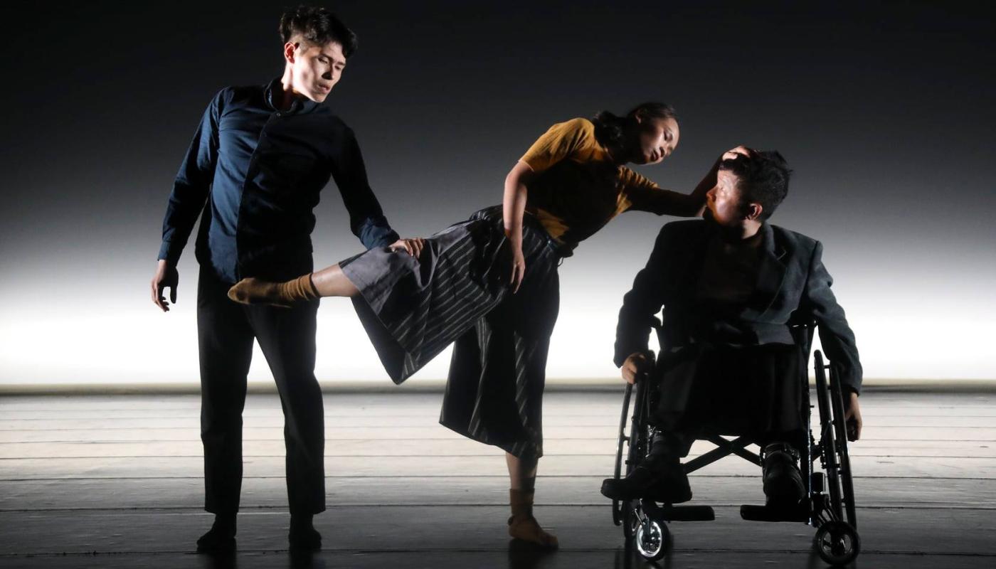 A man standing holding a woman's leg as she leans on a third man in a wheel chair