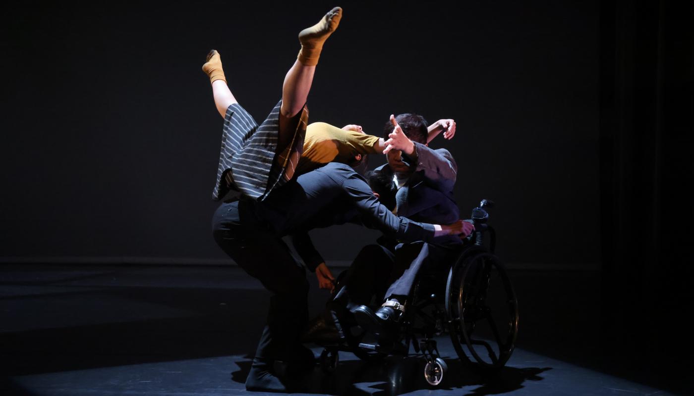 A female dancer balances upside down on the back of a male dancer while also being supported by a dancer in a wheelchair