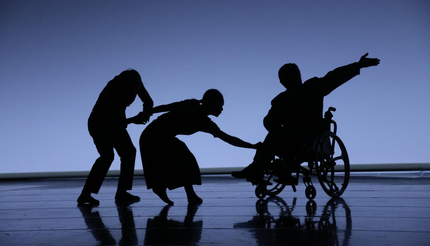 Three silhouetted dancers, two able-bodied and one in a wheelchair, perform on a stage.