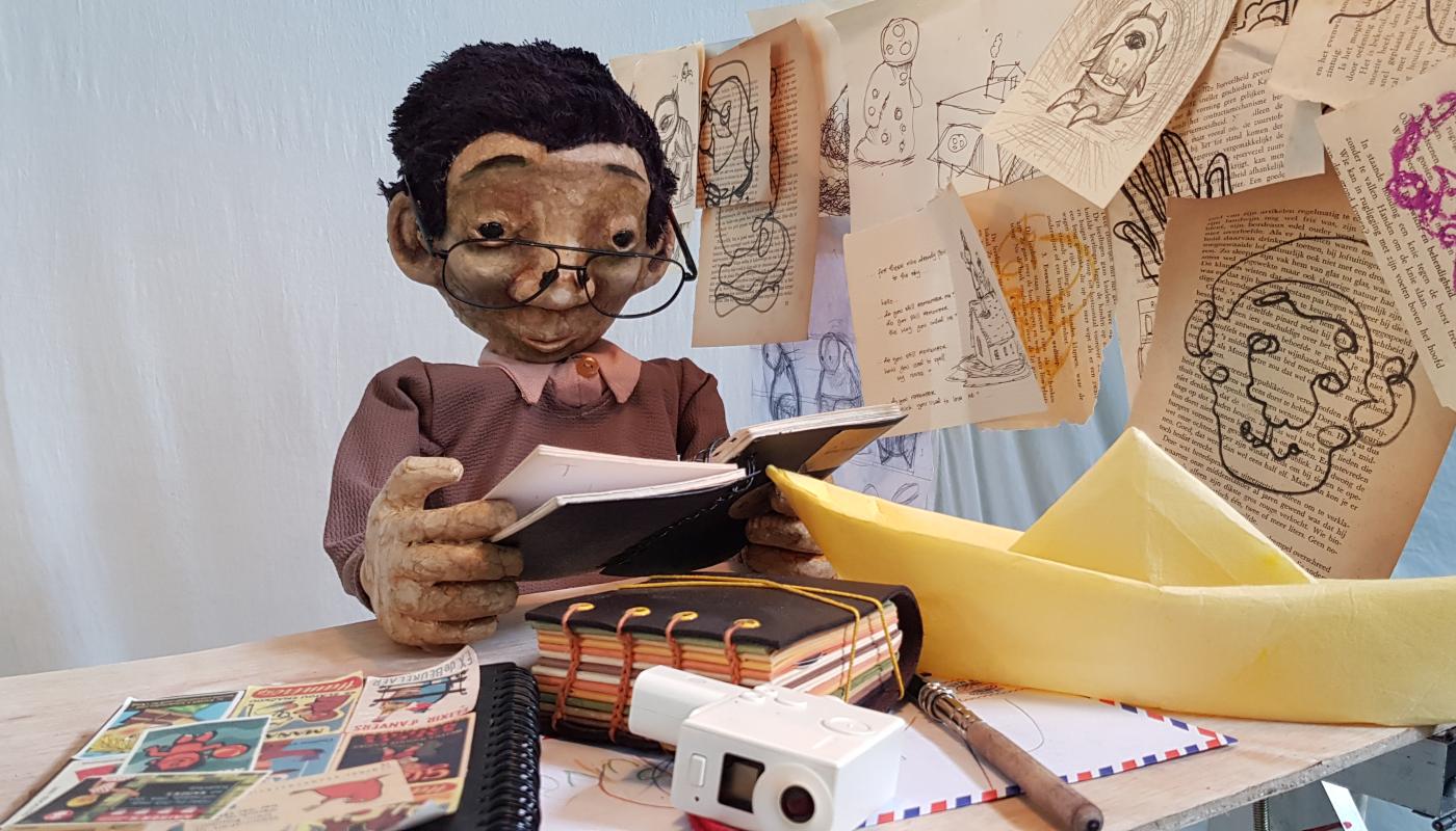 A small puppet sits at a desk and reads a book, surrounded by sketches and papers