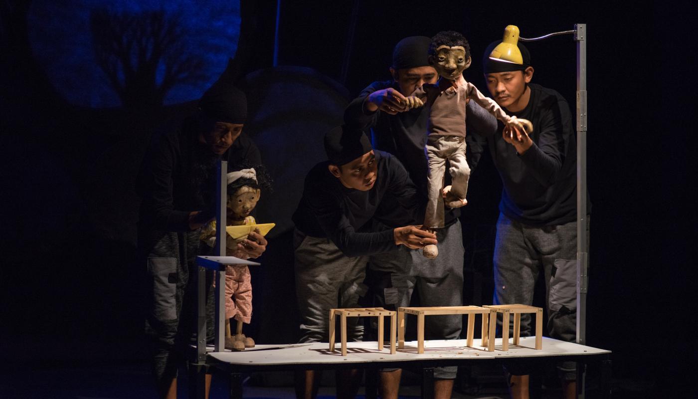 Three puppeteers move a puppet on a small stage with a yellow light above them