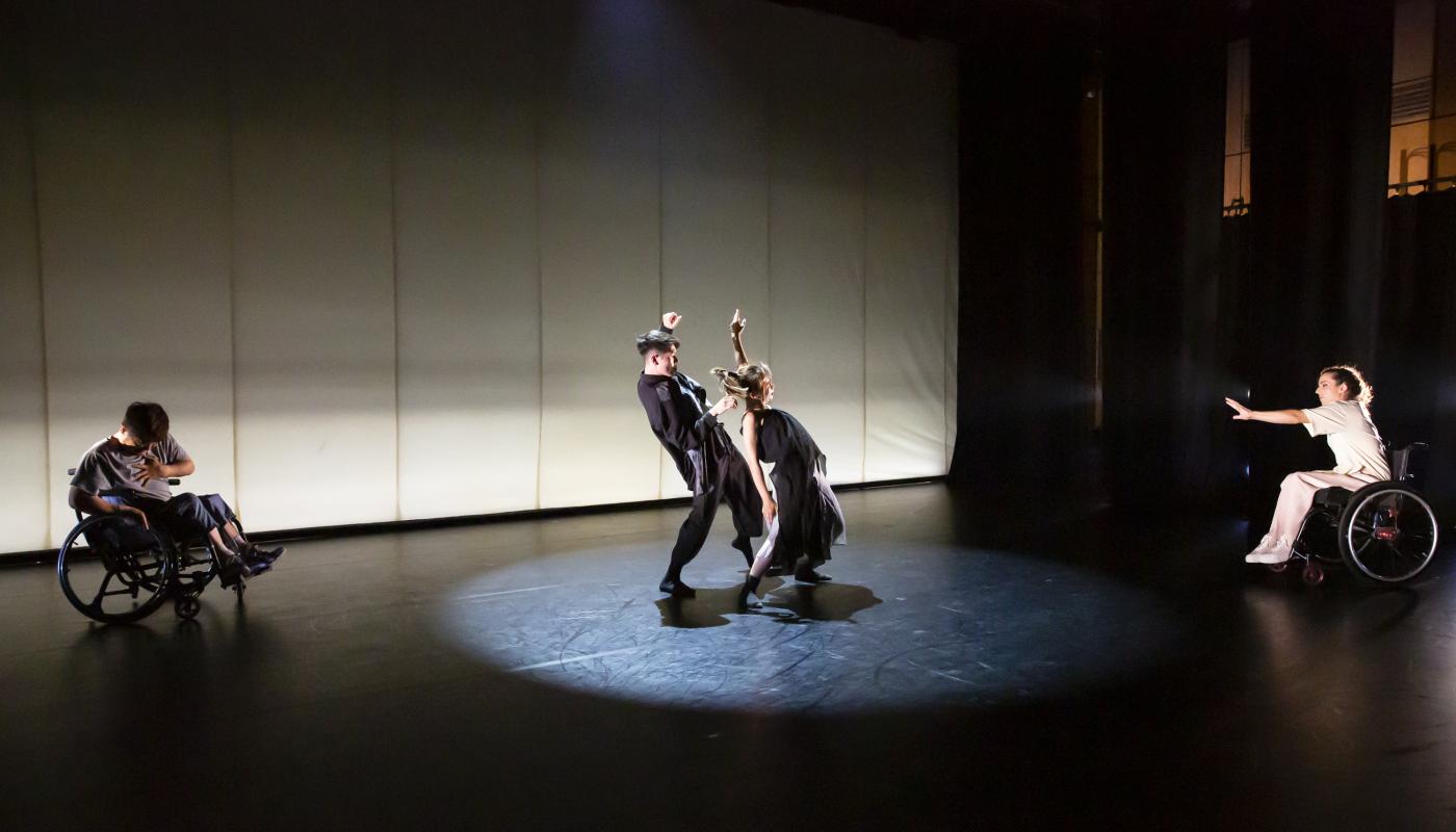 Four dancers, two able-bodied and two in wheelchairs, perform on a spotlighted stage.