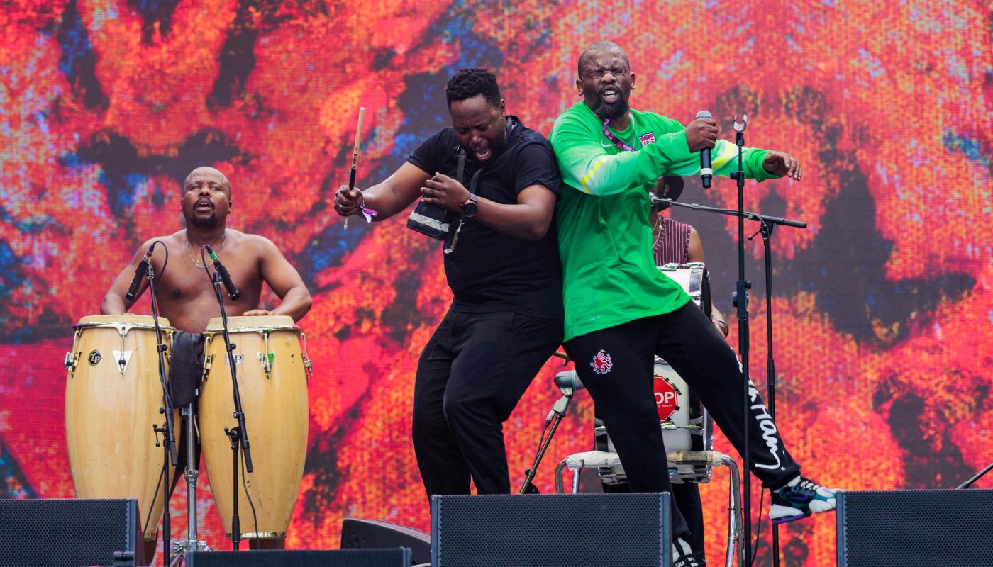 Two men on stage, jumping and bumping into each other, with a drummer behind them