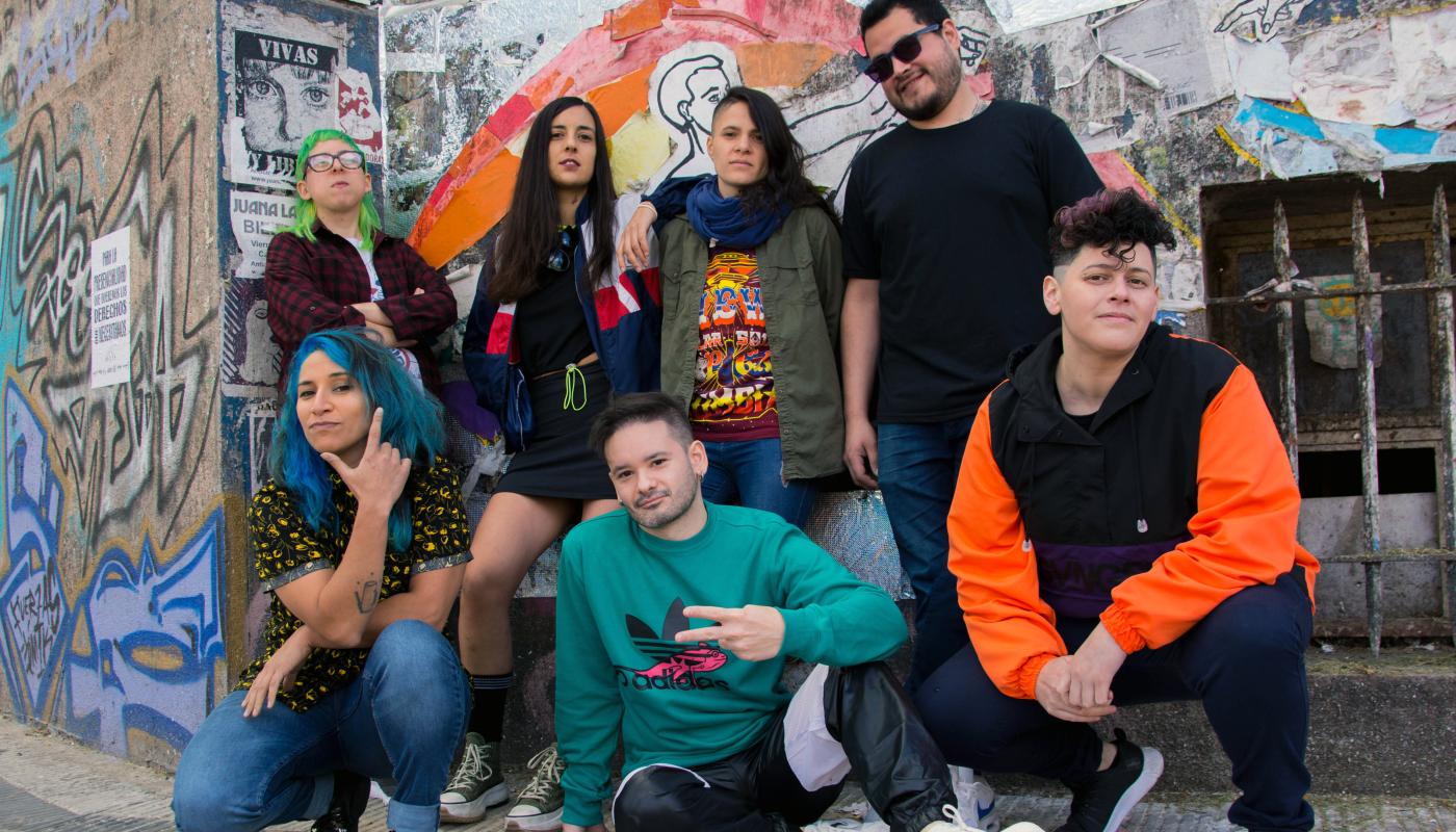 Seven people -- four standing, while three crouch down in front -- pose in front of a graffitied wall