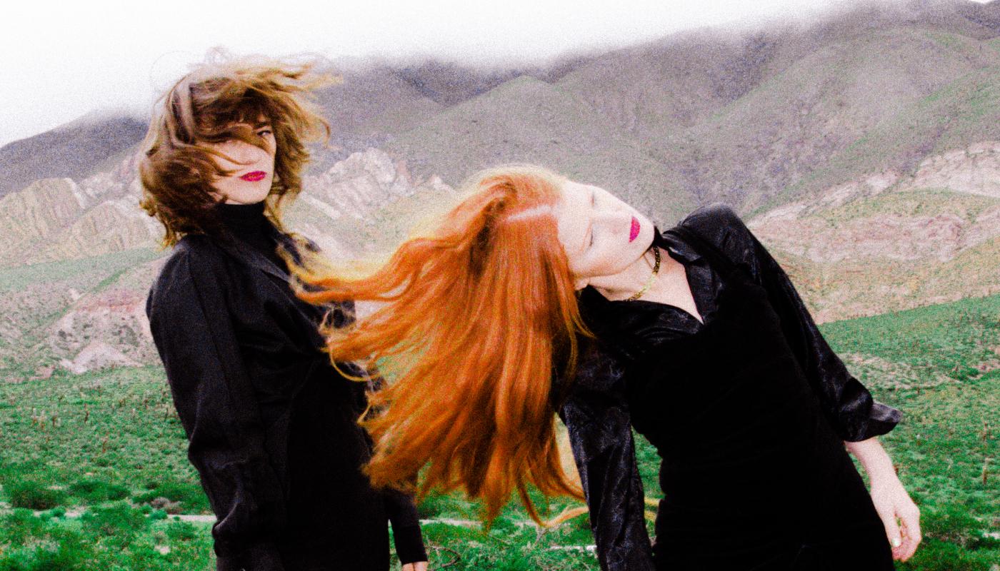 Two women in black, one with dark hair and one with red hair, shake their heads in motion in front of a green landscape. 