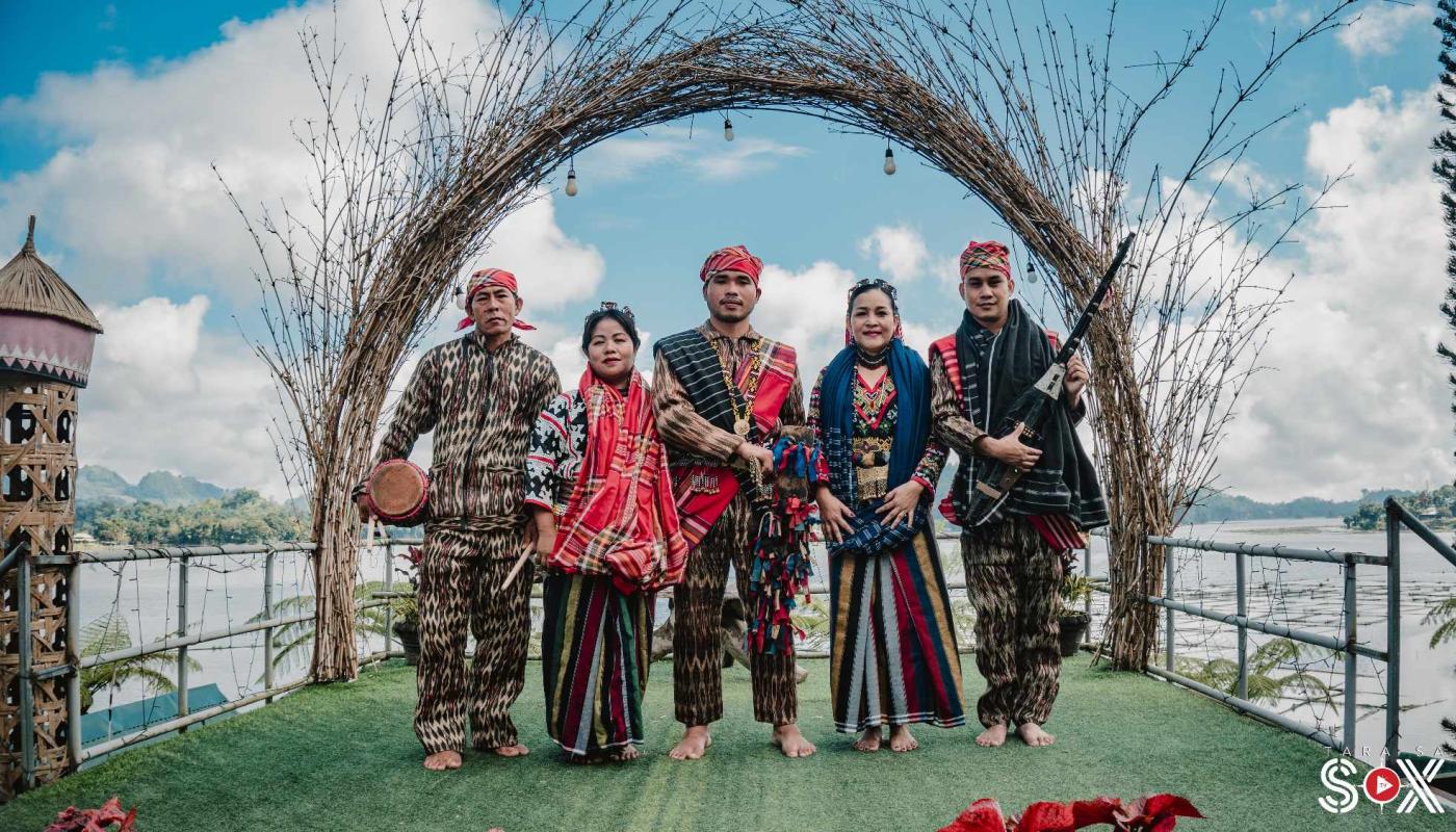 Five indigenous Filipino performers stand in front of a woven archway overlooking a body of water