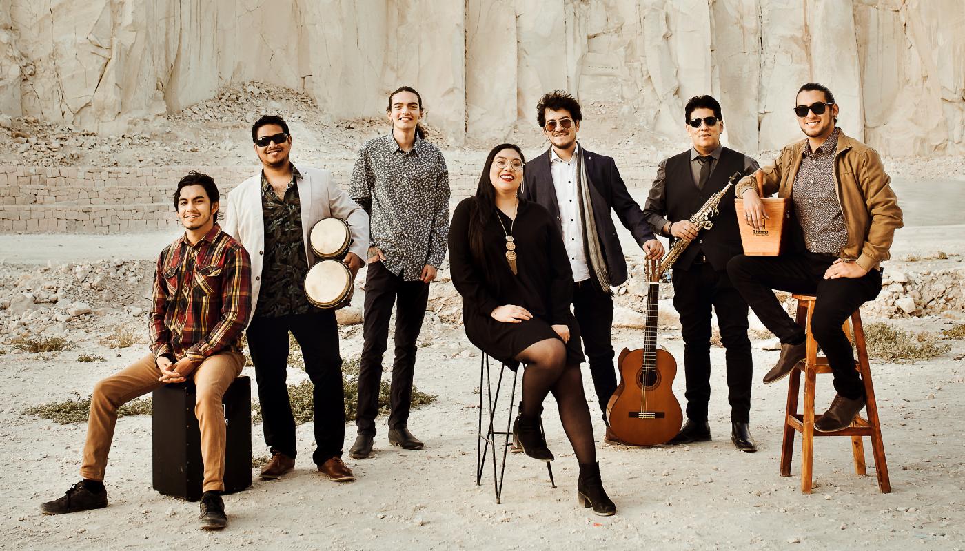 Seven people standing together with various instruments in front of a large white cliff