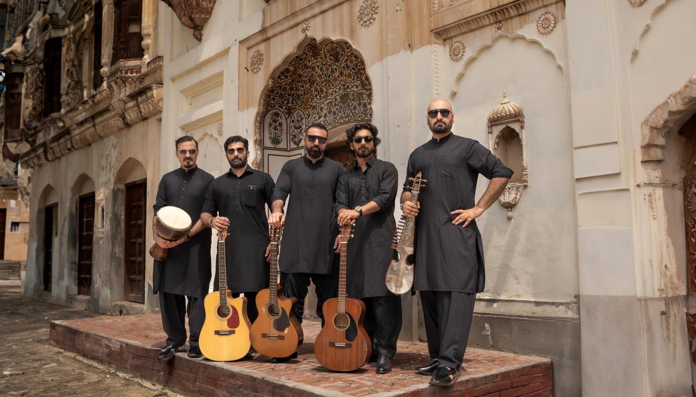 Five men in traditional black outfits standing together with various instruments in front of a historic building in Pakistan