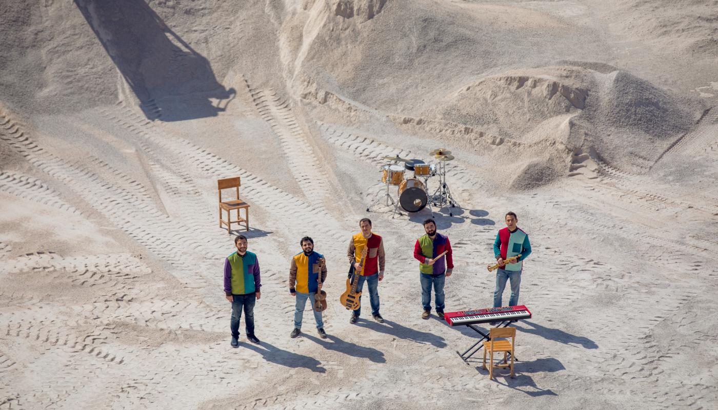 Five musicians in bright colored clothing, holding instruments, standing in a white desert. 