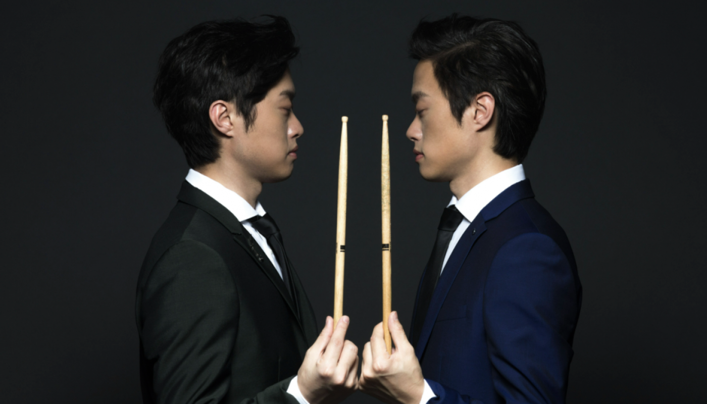 Two men facing each other, each holding a drumstick