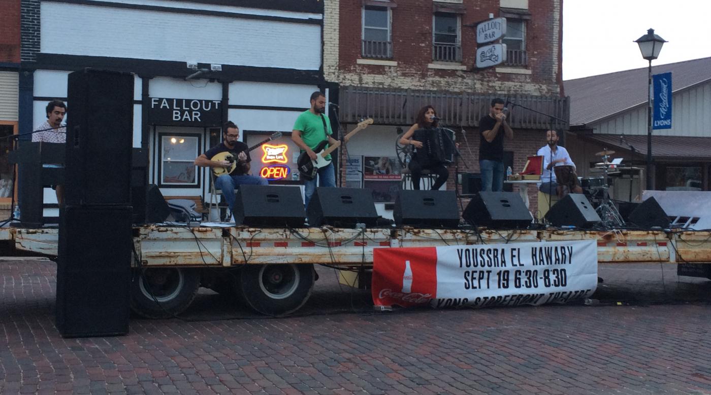 5 musicians perform on a flatbed truck stage. Small midwestern downtown serves as the backdrop.