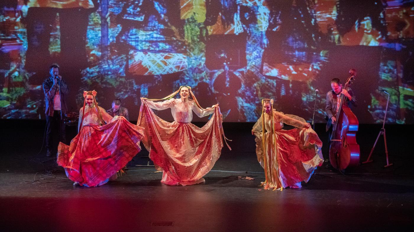 Three women in traditional Ukrainian dress perform with 3 musicians in the background and a colorful backdrop. 