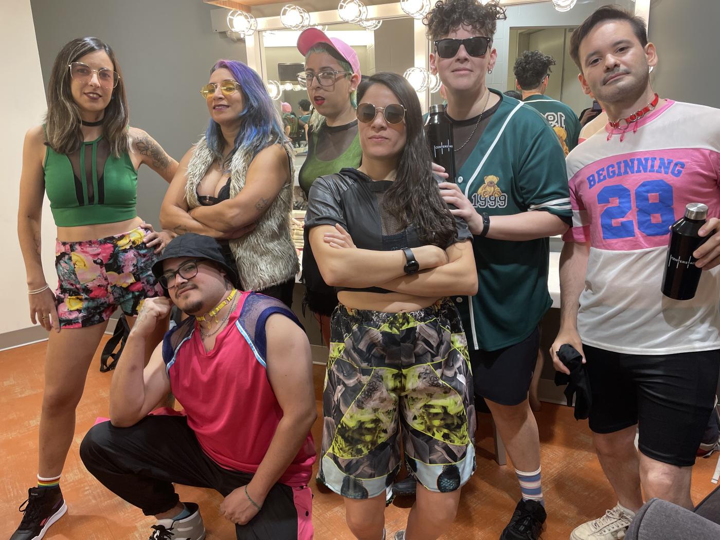 A band of seven musicians wearing colorful street clothes pose backstage