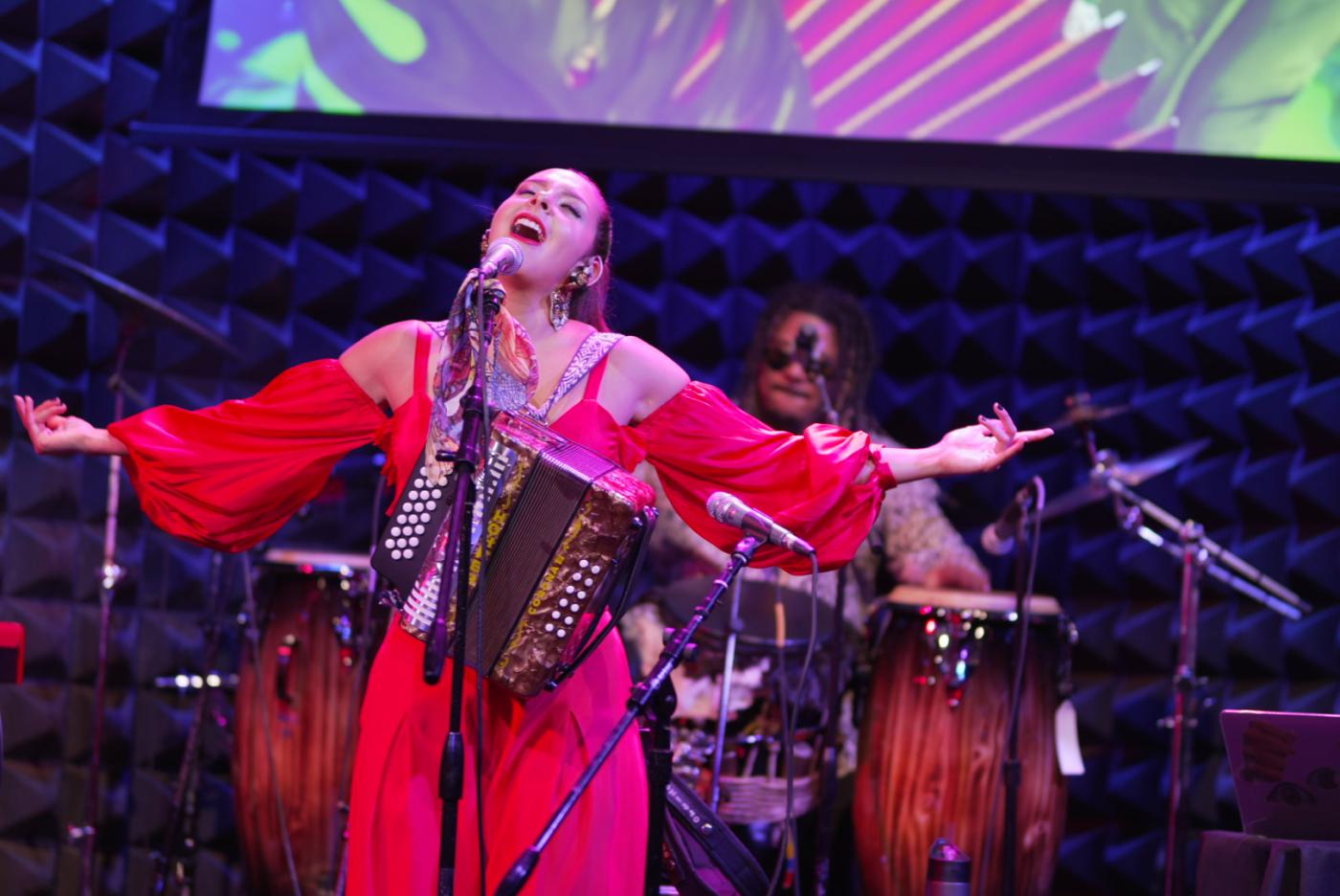 A woman in a red dress singing on a stage, arms outstretched, with an accordion