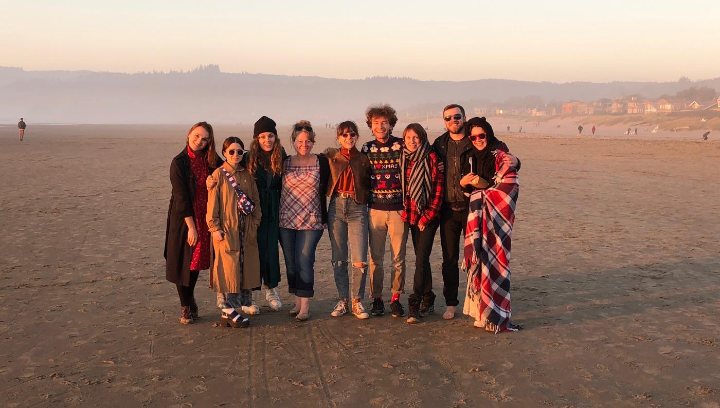 Nine people dressed for Pacific Northwest weather standing arm-in-arm on a sunny and misty beach at low tide. Mountains, trees, and houses create the background.