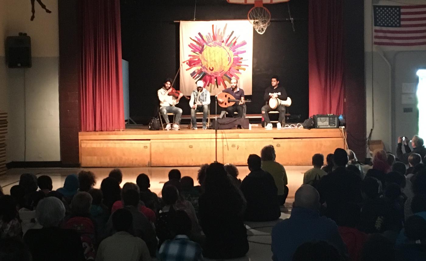 A 4 piece band plays classical Egyptian instruments on a stage in a school theater/gymnasium. An audience of students fill the foreground