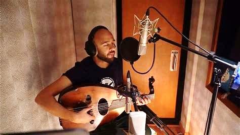 Mohamed Abozekry in Recording Booth, Photo Courtesy of Mohamed Abozekry A man in a black T-shirt is playing an oud in a recording booth and singing into a microphone while wearing headphones