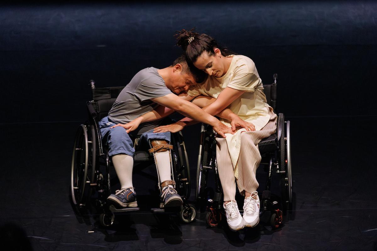 Two dancers, using wheelchairs, embrace on stage