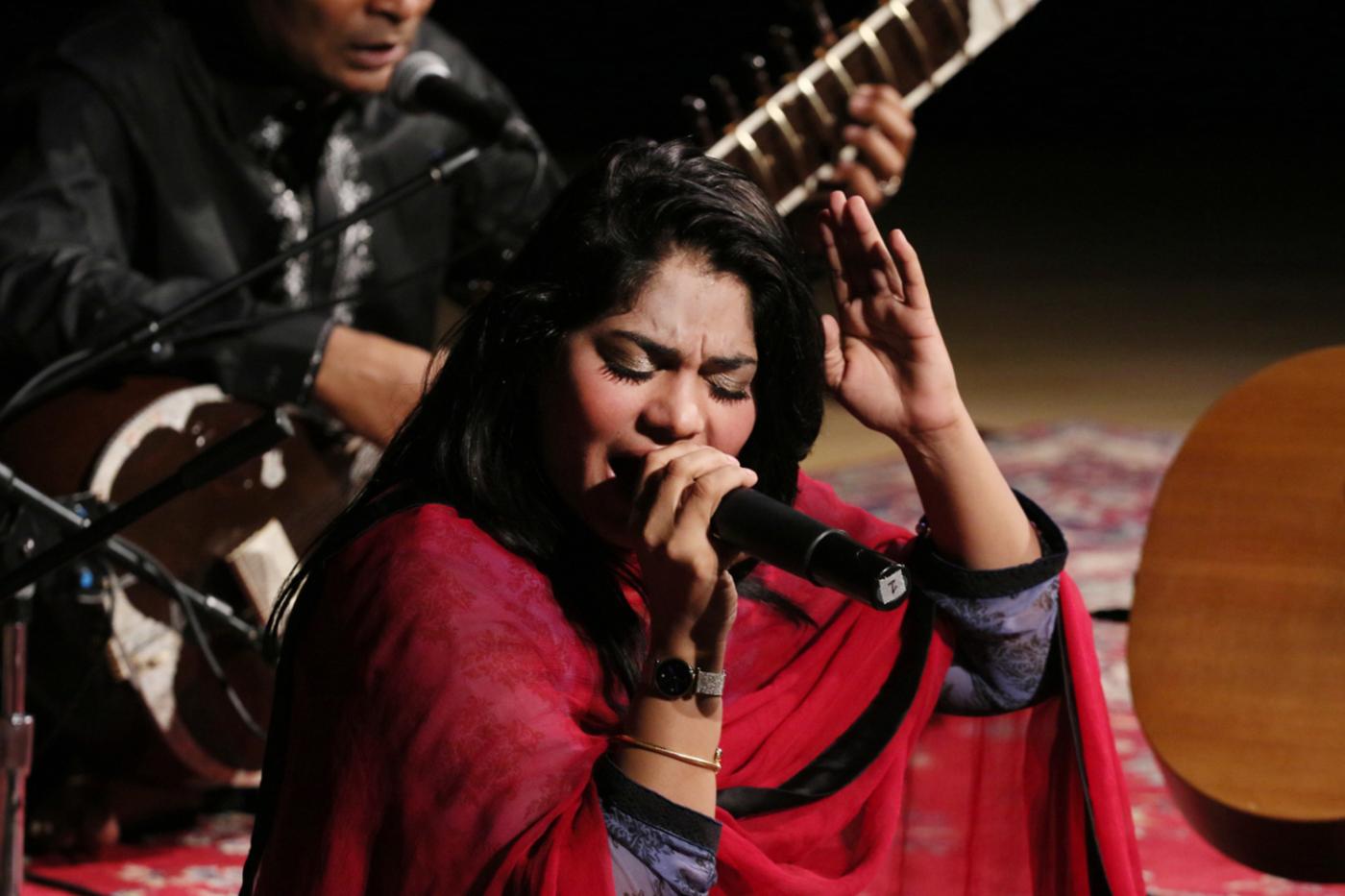 Woman sitting down on stage, singing with her hand in the air, wearing a red sari