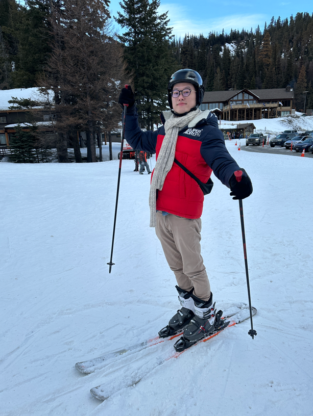 A person in a red and black jacket waves while standing on skiis