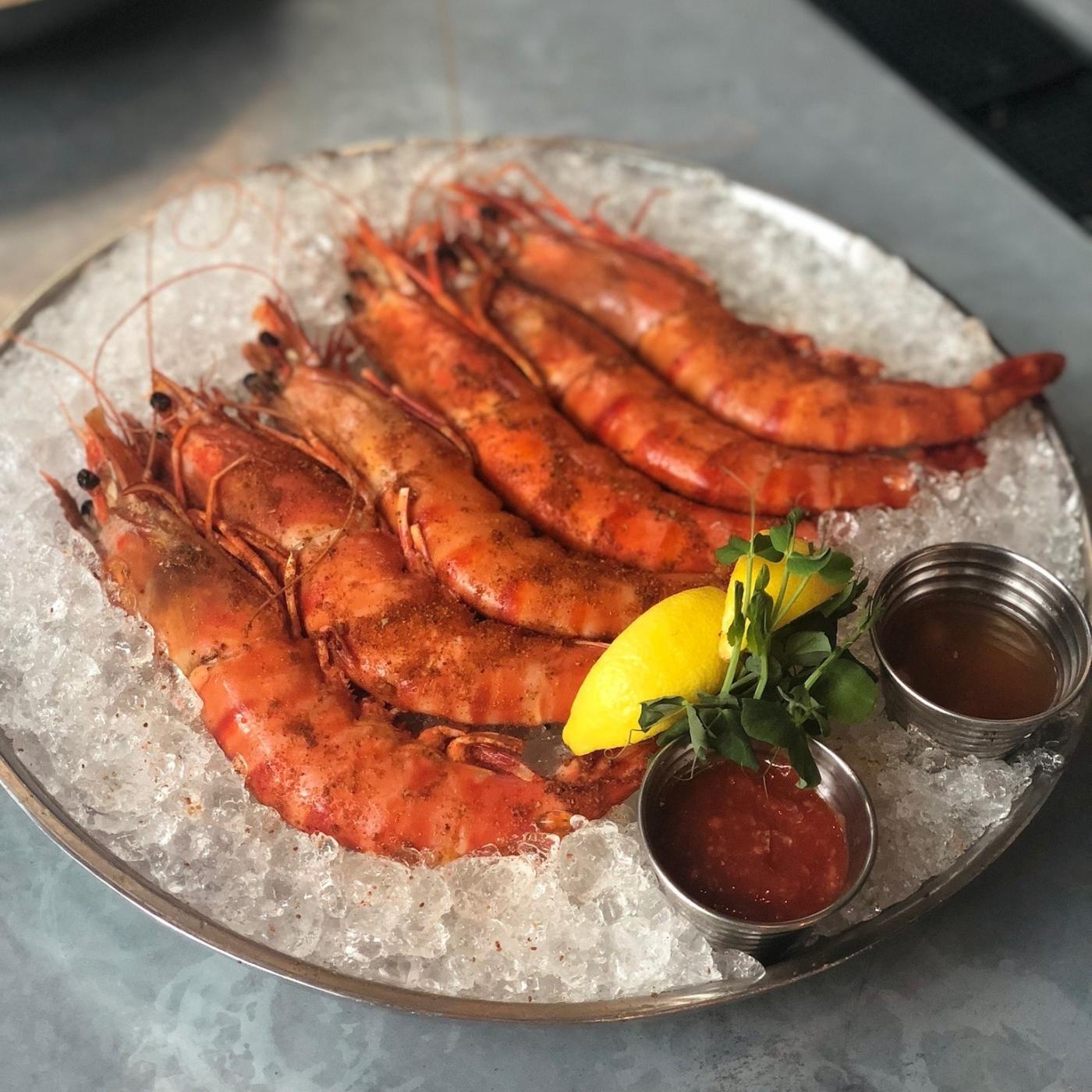 A photo of shrimp laying on a bed of ice with lemon, herbs, and sauces