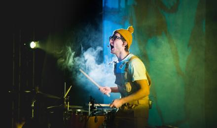 A man playing drums wearing a yellow hat, silhoutted by a spotlight