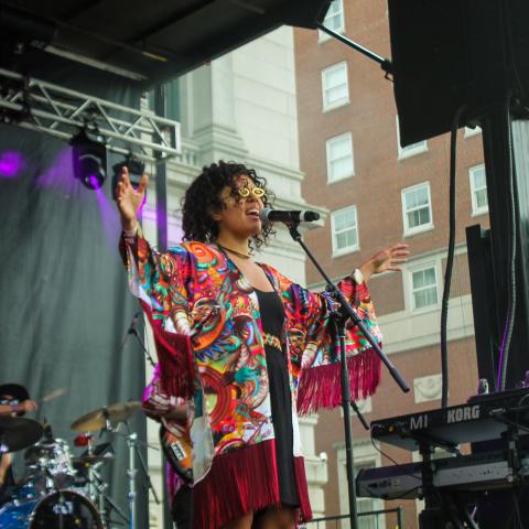 A woman in a colorful shawl sings with her hands outstretched on an outdoor stage.