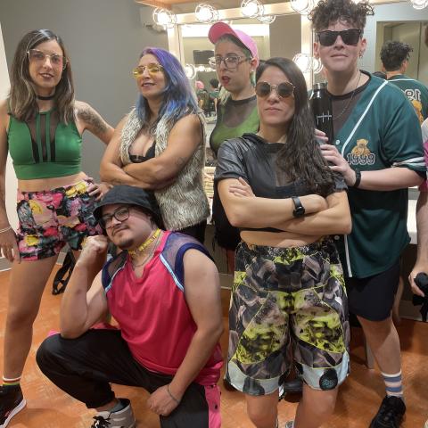 A band of seven musicians wearing colorful street clothes pose backstage