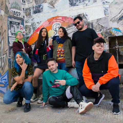 Seven people stand and crouch in front of a graffitied wall.