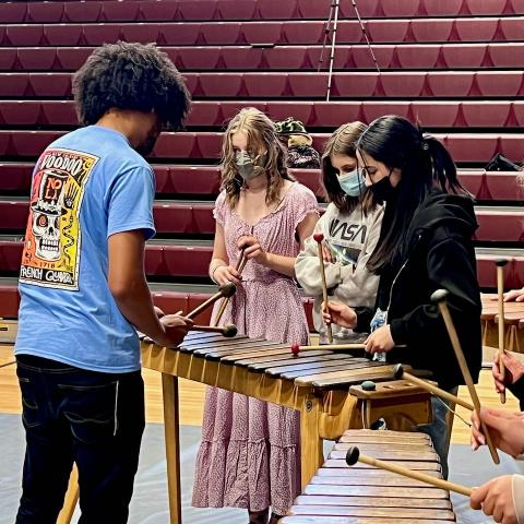 A man with an afro in a blue shirt instructs three students how to play the marimba