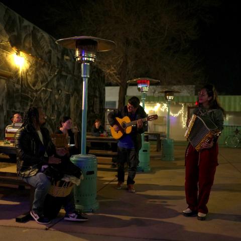 Several musicians gather outside at night playing instruments next to a heat lamp.