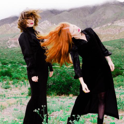 Two women in black, one with dark hair and one with red hair, shake their heads in motion in front of a green landscape. 