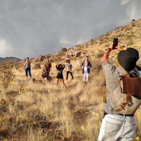 A group of musicians pose in a desert landscape while a man takes a photo of them with his phone.