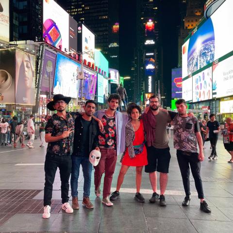 A group of six adults pose in the middle of Time Square New York at night with many billboards illuminated in the background.