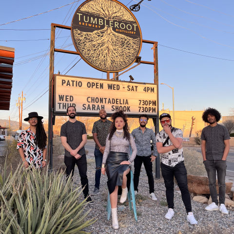 A group of musicians stand in a desert setting in front of a sign that reads "Tumbleroot Brewery Distillery"