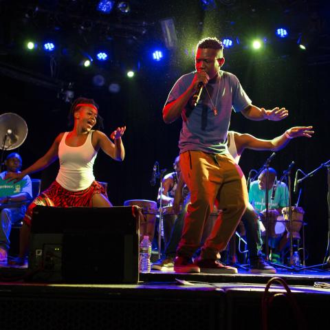 People performing on stage, singing into a microphone and someone dancing in the background