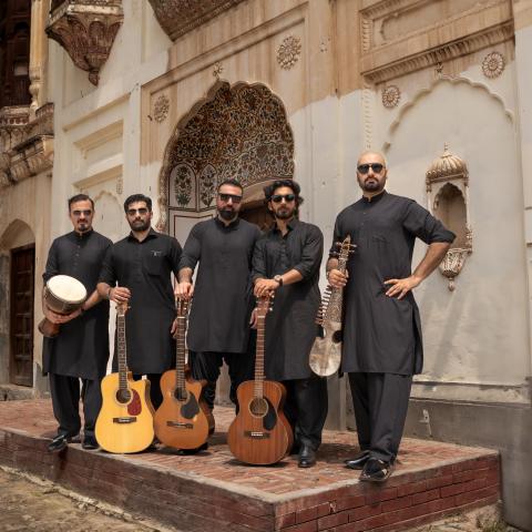 Five men in traditional black outfits standing together with various instruments in front of a historic building in Pakistan