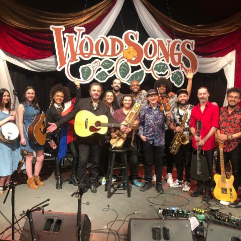 A large group of musicians, some holding various string instruments, pose on a stage with red curtains and a sign that reads "WoodSongs"