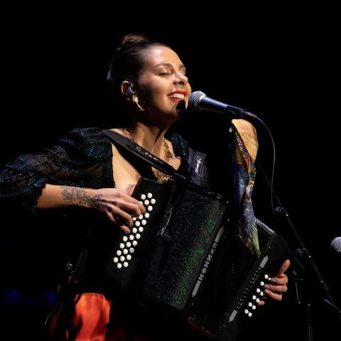 A woman spotlighted on a dark stage sings and plays accordion